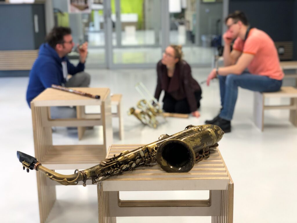 Ein Saxophon liegt im Vordergrund und dahinter sitzen drei Personen die sich unterhalten