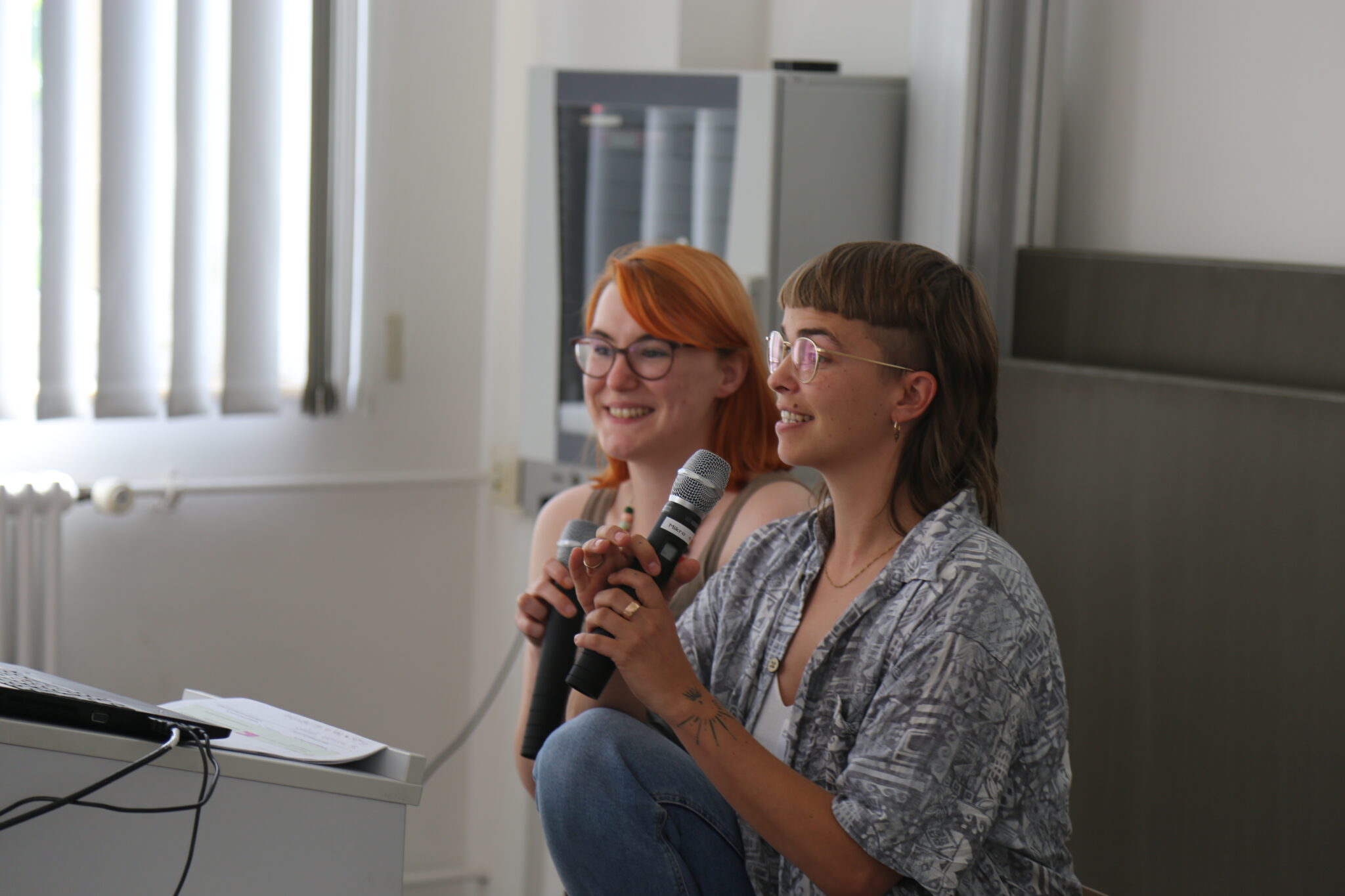 Zwei Personen sitzen vor einem Bildschirm und sprechen in Mikros