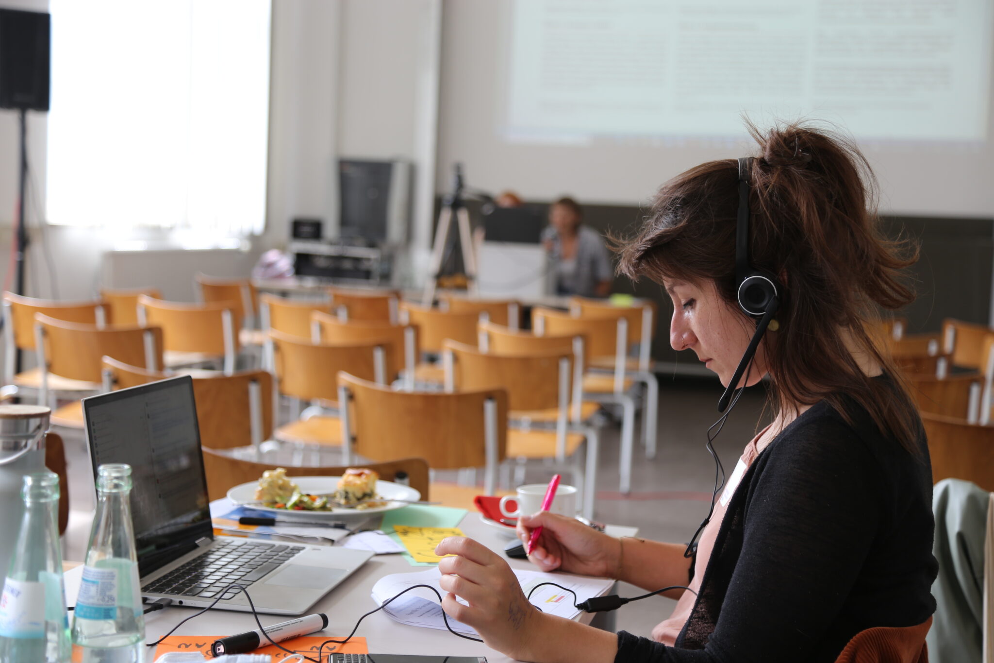 Eine Frau sitzt mit einem Headset vor einem Laptop und schaut auf ein Blatt Papier